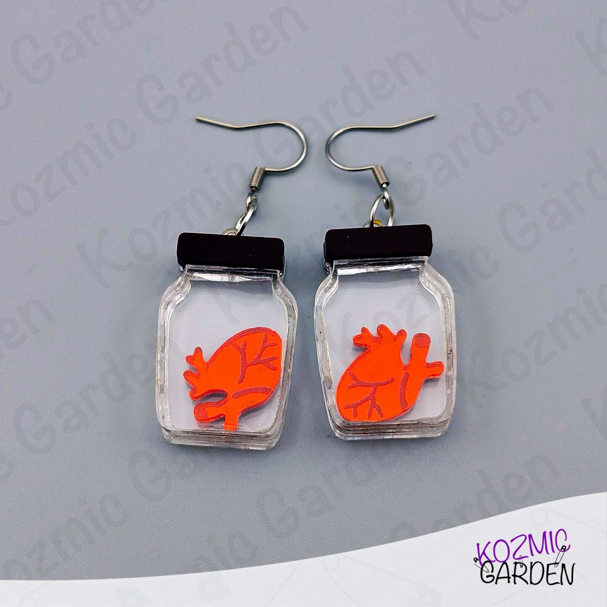 Earrings with red anatomical heart design in clear jars on a gray background, branded 'Kozmic Garden'.