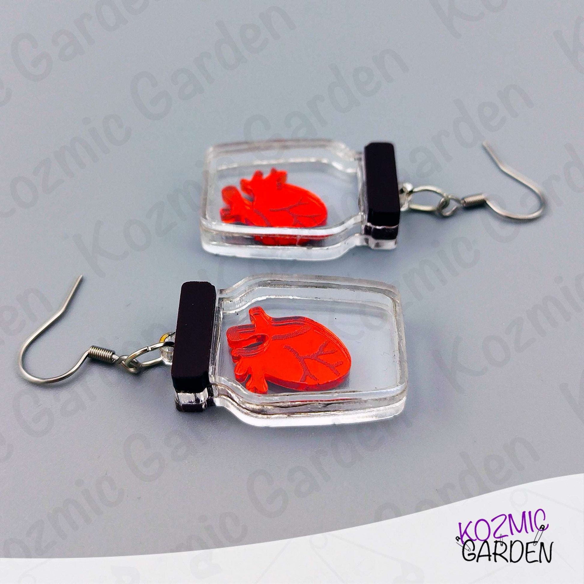 Earrings with red heart design in clear square frames on a gray background, branded 'Kozmic Garden'.