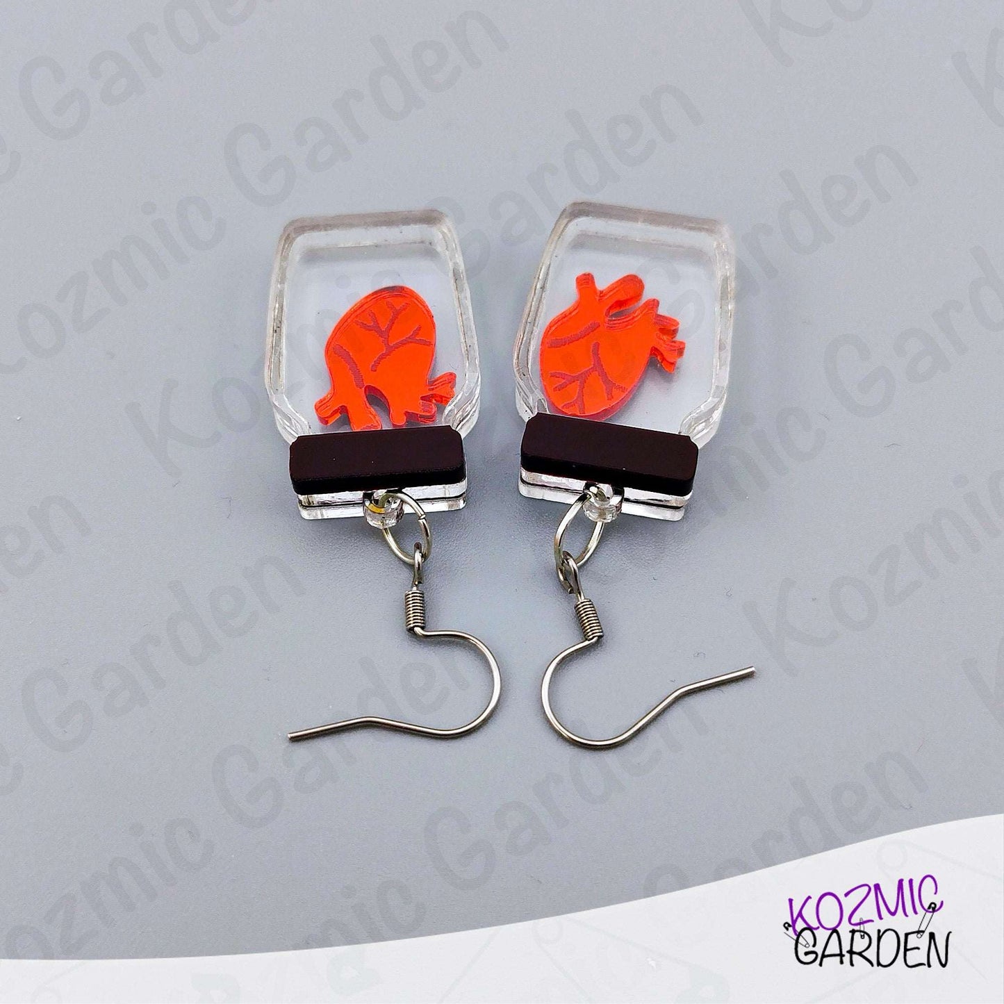 Earrings with red heart design in clear rectangular frames on a gray background, branded 'Kozmic Garden'.