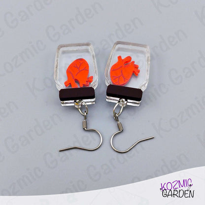 Earrings with red heart design in clear rectangular frames on a gray background, branded 'Kozmic Garden'.