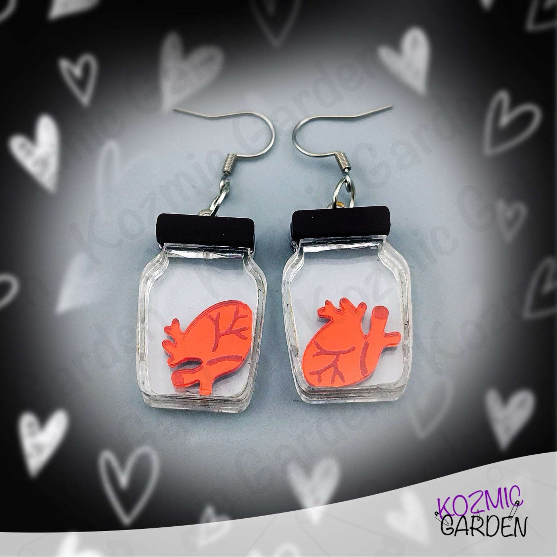 Earrings with red heart design in clear jars on a heart-patterned background, branded 'Kozmic Garden'.