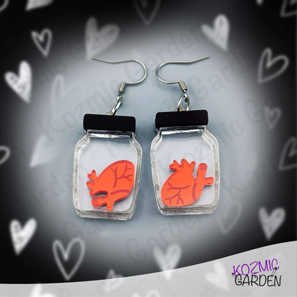 Earrings with red heart design in clear jars on a heart-patterned background, branded 'Kozmic Garden'.