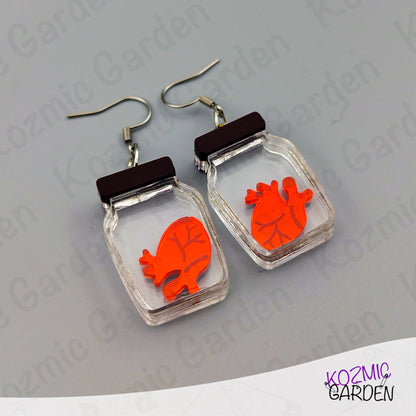 Earrings with orange heart design in clear rectangular frames on a gray background, branded 'Kozmic Garden'.