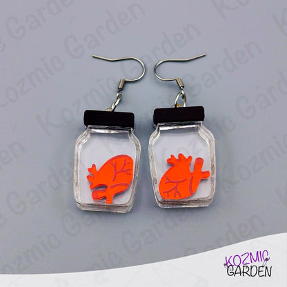 Earrings with red anatomical heart design in clear jars on a gray background, branded 'Kozmic Garden'.