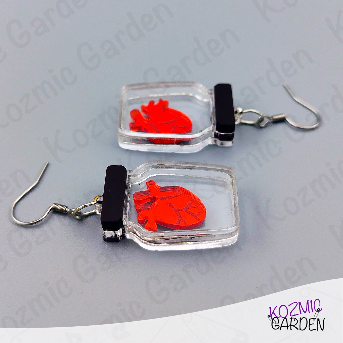 Earrings with red heart design in clear square frames on a gray background, branded 'Kozmic Garden'.