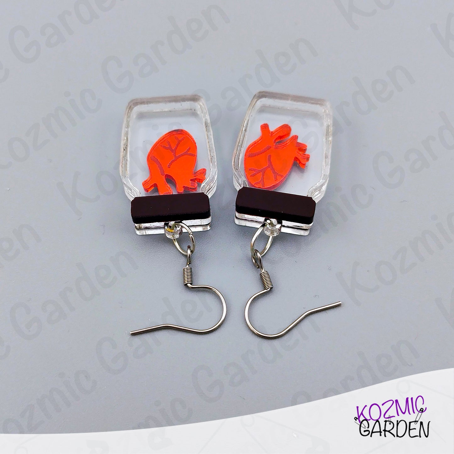 Earrings with orange heart design in clear rectangular frames on a gray background, branded 'Kozmic Garden'.