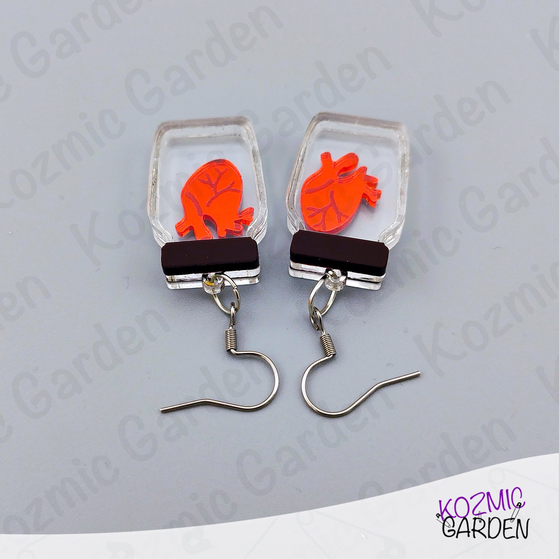 Earrings with orange heart design in clear rectangular frames on a gray background, branded 'Kozmic Garden'.
