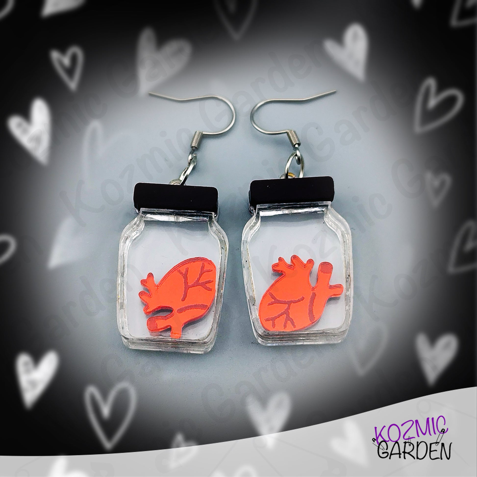 Earrings with red heart design in clear jars on a heart-patterned background, branded 'Kozmic Garden'.