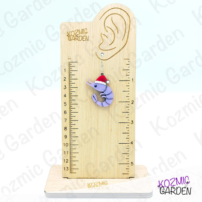 Wooden ear gauge with a ruler and a purple earring shaped like a shrimp with a Santa hat on a white background.