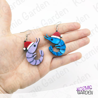 Hand holding two novelty earrings shaped like shrimps with Santa hats on a white background.