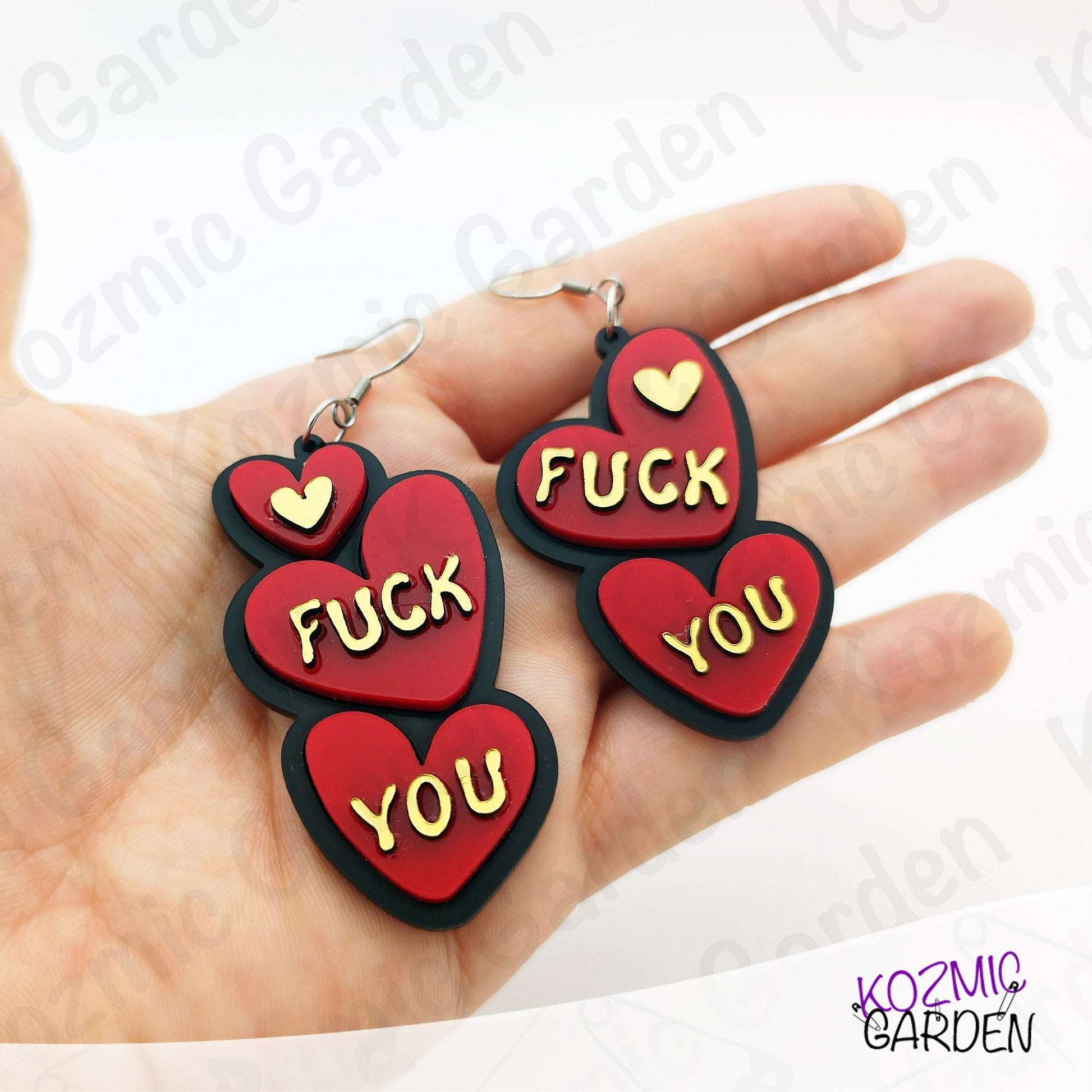 Red heart-shaped earrings with 'FUCK YOU' text held in a hand, brand 'Kozmic Garden' visible.