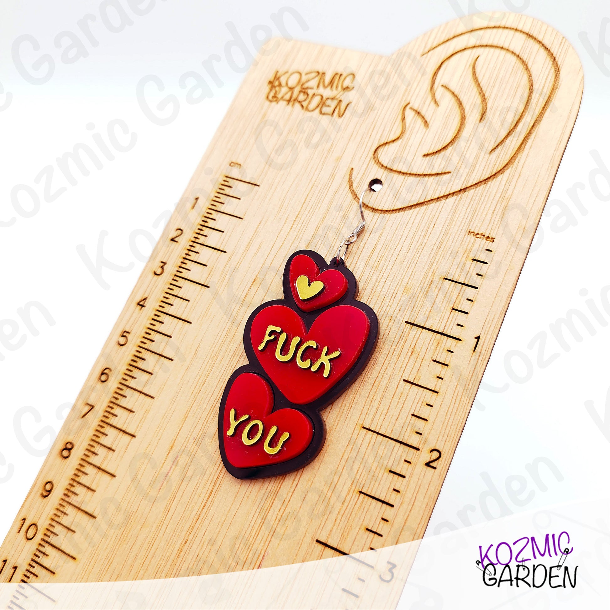 Red heart-shaped earrings with gold text on a wooden ruler, branded 'Kozmic Garden'.