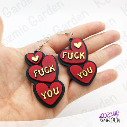 Red heart-shaped earrings with 'FUCK YOU' text held in a hand, brand 'Kozmic Garden' visible.