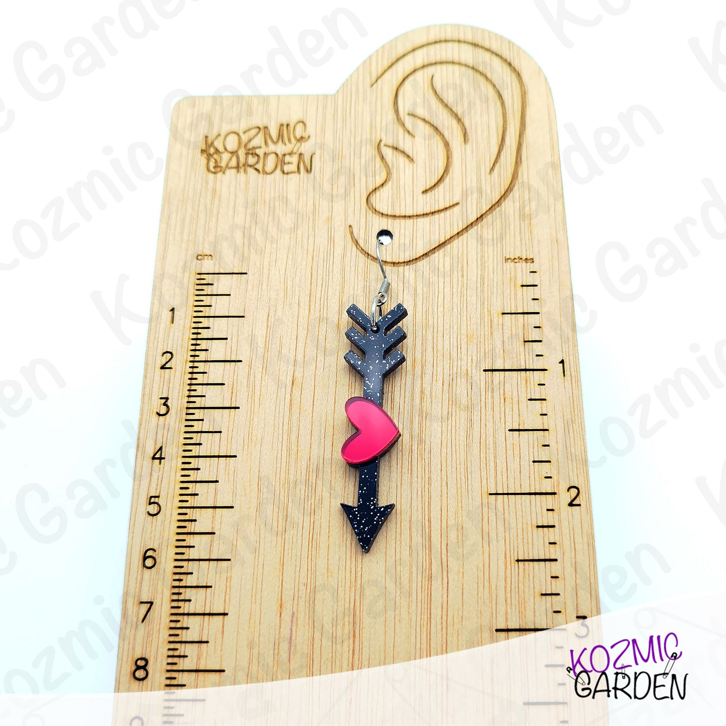 Earring with a heart and arrow design on a wooden ruler for scale, branded 'Kozmic Garden'.