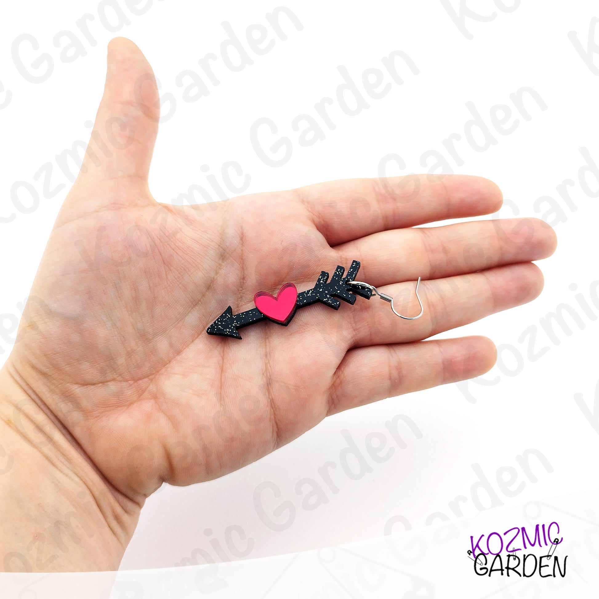 Hand holding a small black arrow-shaped earring with a pink heart on a white background.