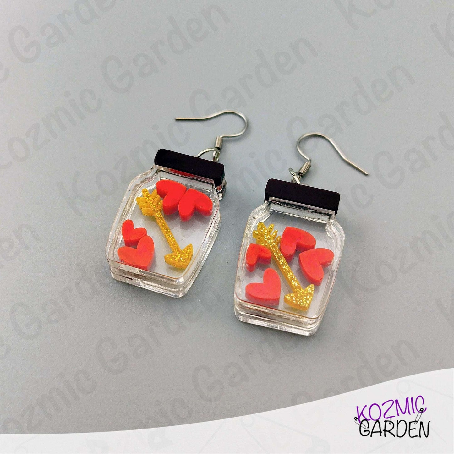 Earrings with heart and arrow design in clear jars on a gray background with 'Kozmic Garden' branding.