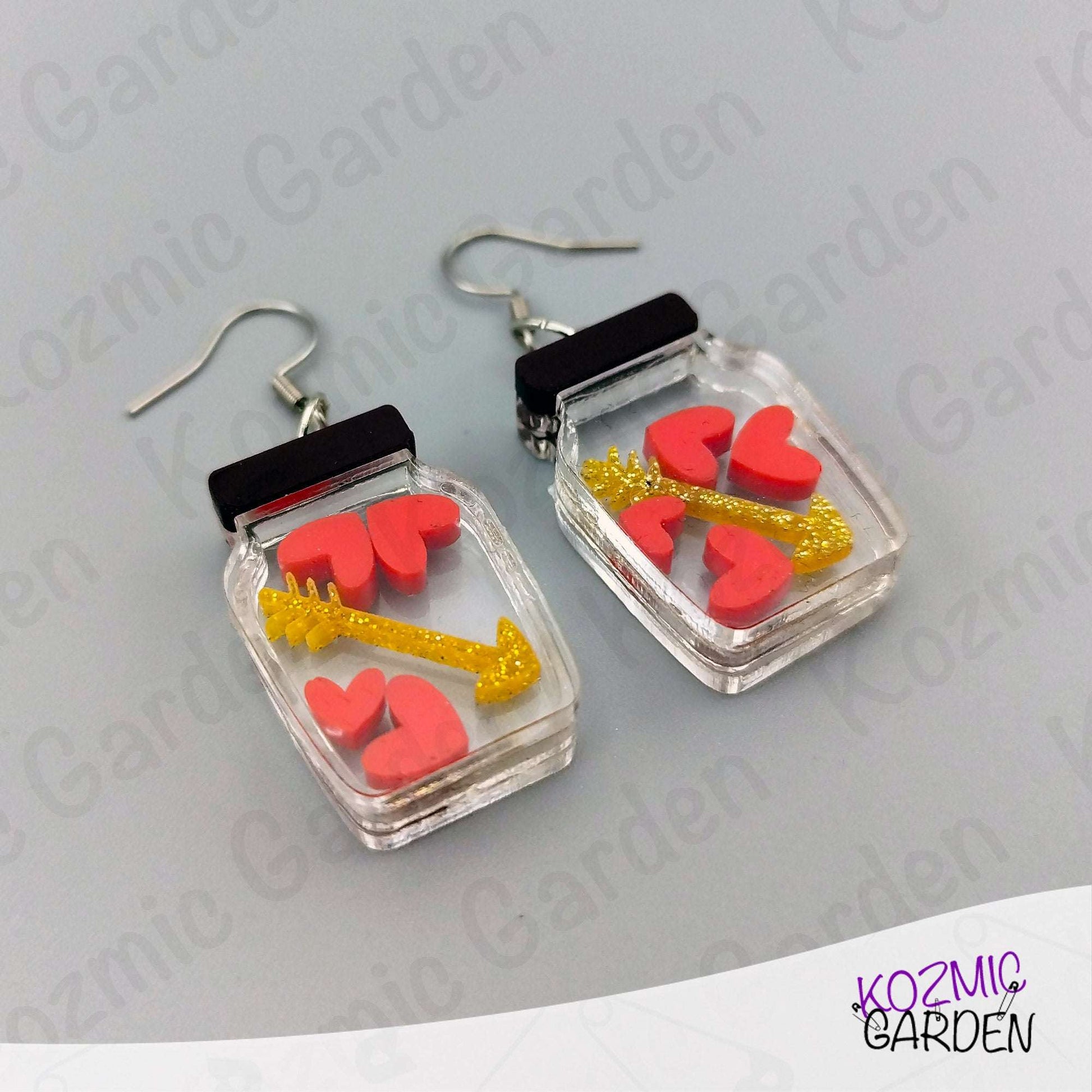Earrings with heart and arrow design in clear case on gray background, brand 'Kozmic Garden' visible.