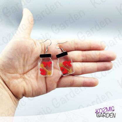 Hand holding earrings with clear jars containing red hearts and golden arrow, brand 'Kozmic Garden' visible.