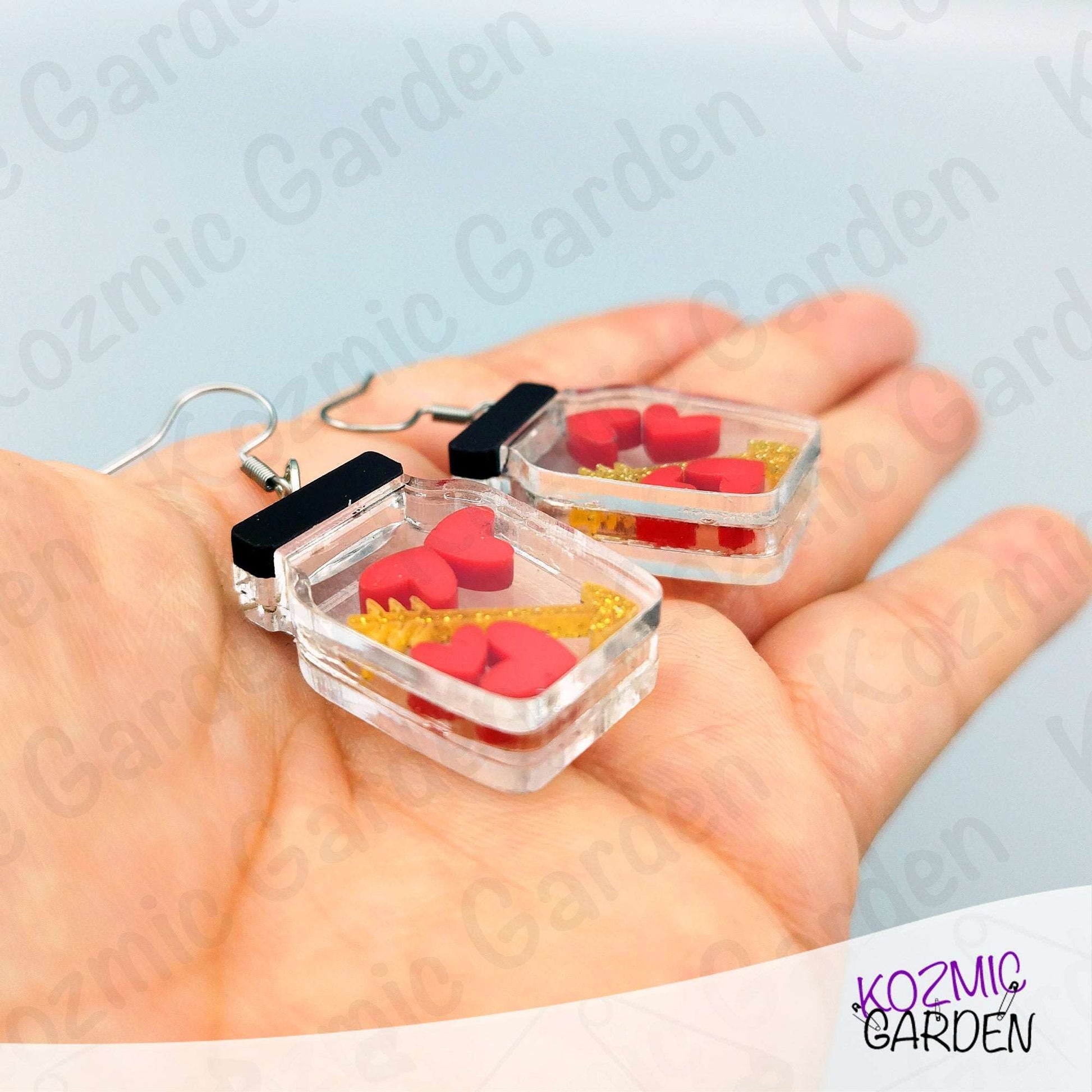 Clear heart-shaped earrings with red and yellow arrow held in a hand against a light background with 'Kozmic Garden' branding.