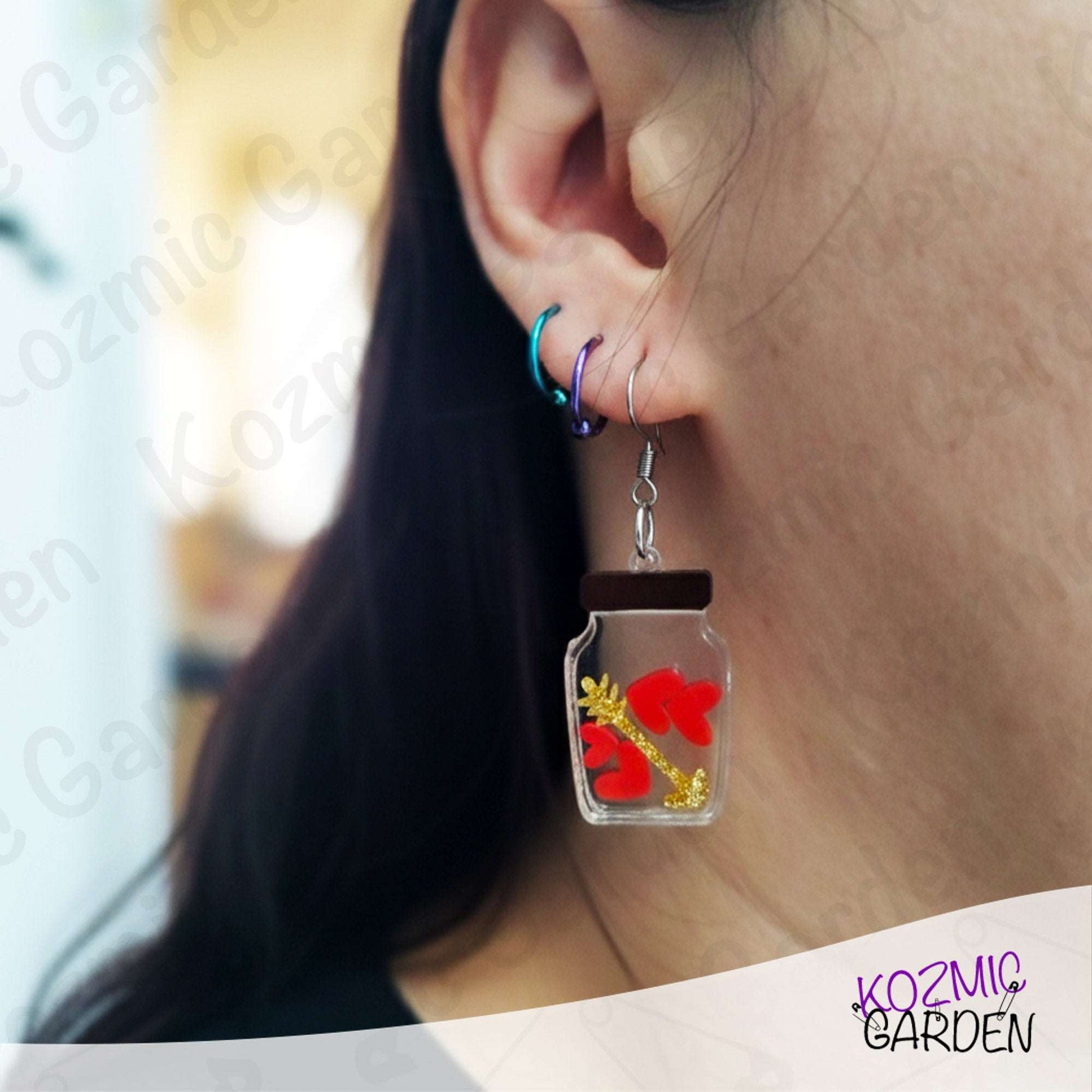 Earring with a jar design featuring hearts and an arrow worn by a person.