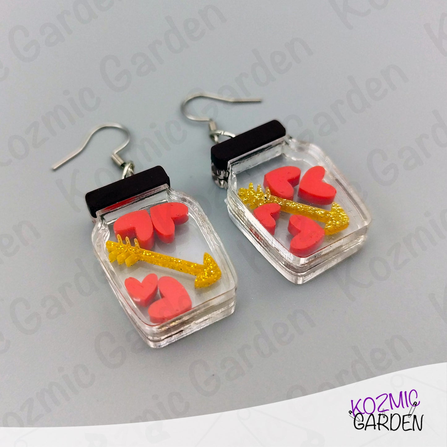 Earrings with heart and arrow design in clear case on gray background, brand 'Kozmic Garden' visible.