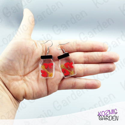 Hand holding earrings with clear jars containing red hearts and golden arrow, brand 'Kozmic Garden' visible.