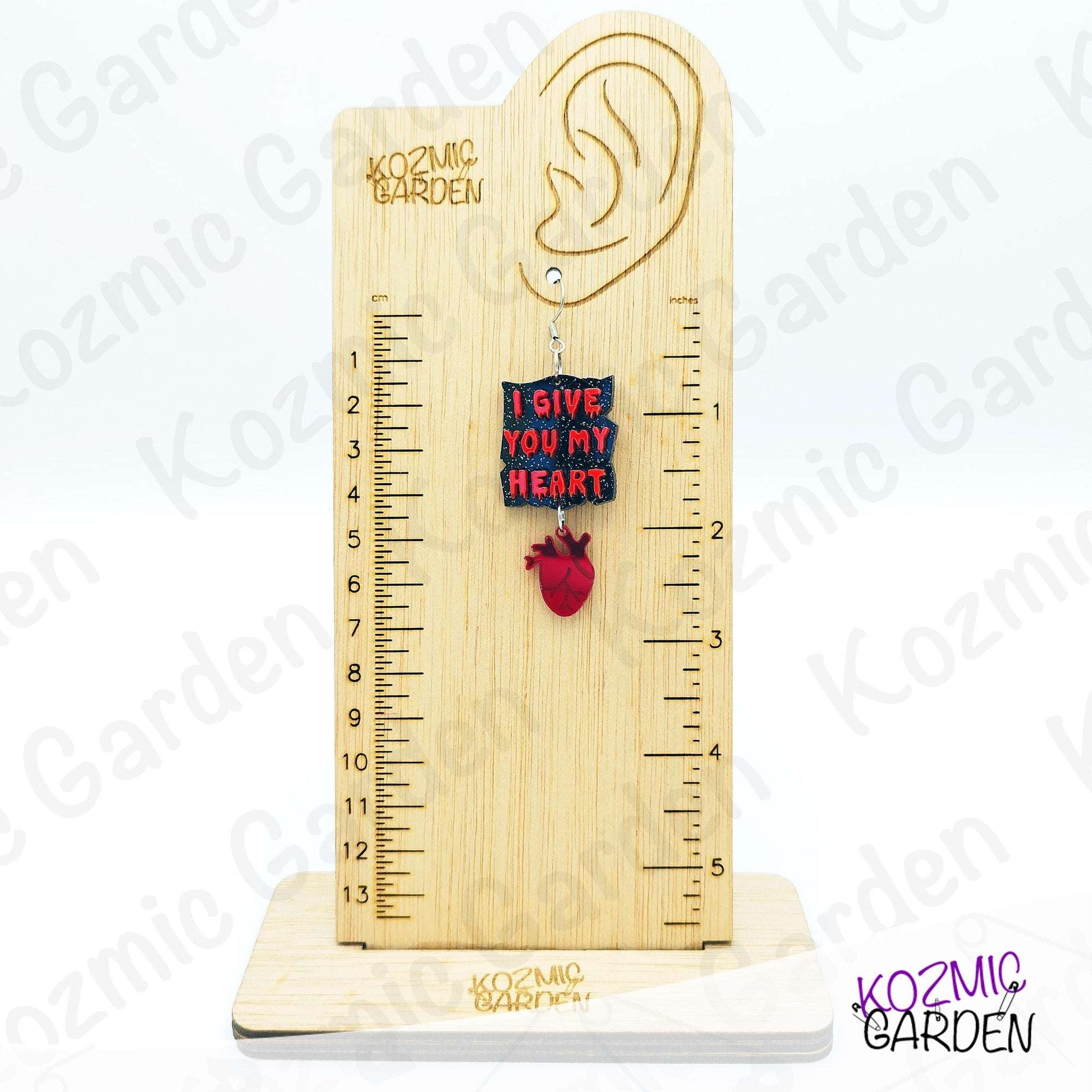 Wooden ear gauge with heart-shaped pendant and ruler on a white background, branded 'Kozmic Garden'.