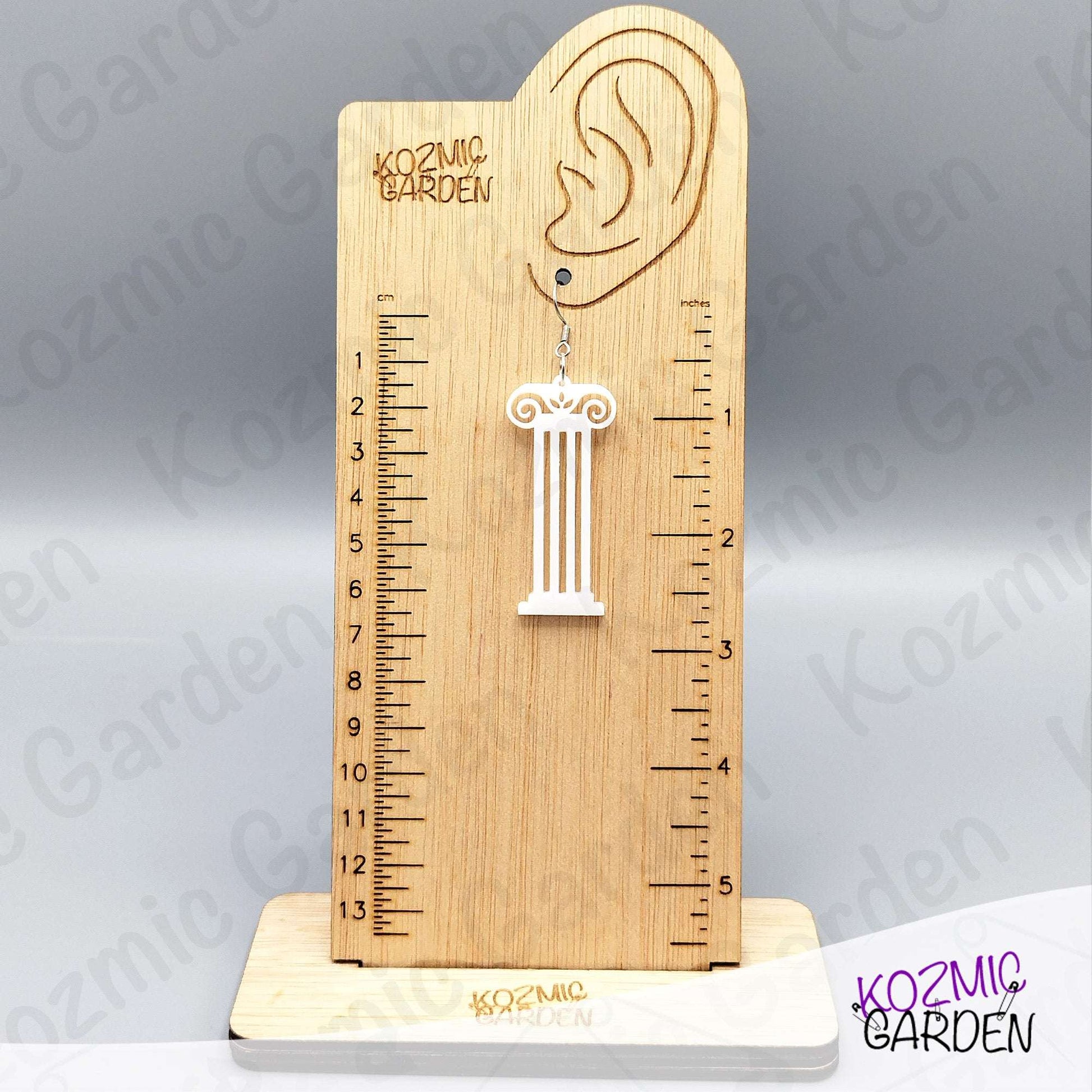 Wooden ruler with an ear-shaped hole and a column-shaped earring on a gray background with 'Kozmic Garden' branding.