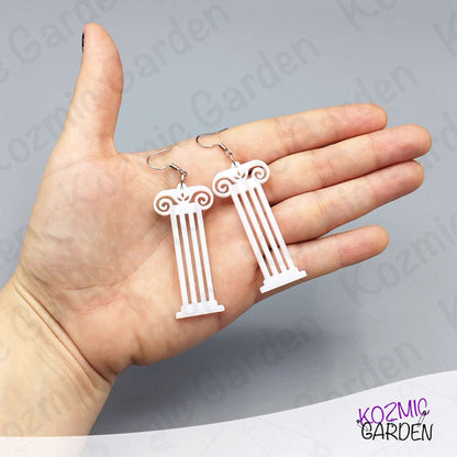 White column-shaped earrings held in a hand with a gray background and 'Kozmic Garden' branding.