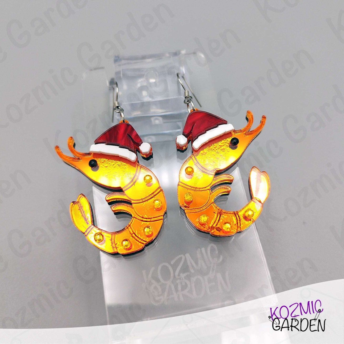 Shrimp-shaped earrings with Santa hats on a Kozmic Garden branded stand.
