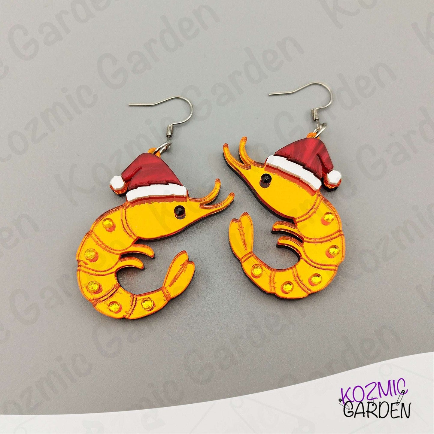 Shrimp-shaped earrings with Santa hats on a gray background, branded 'Kozmic Garden'.