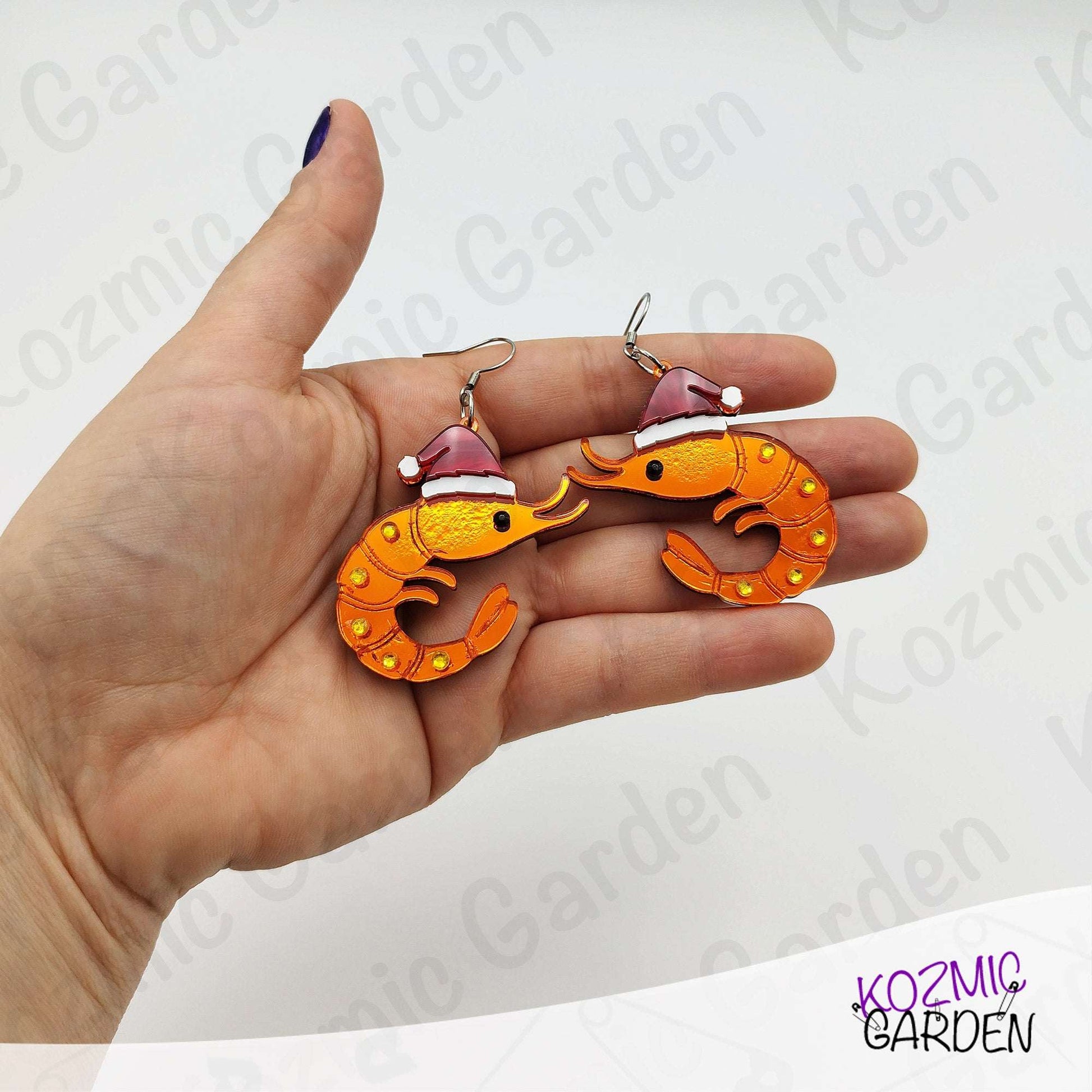 Hand holding two orange seahorse-shaped earrings with red hats against a white background.