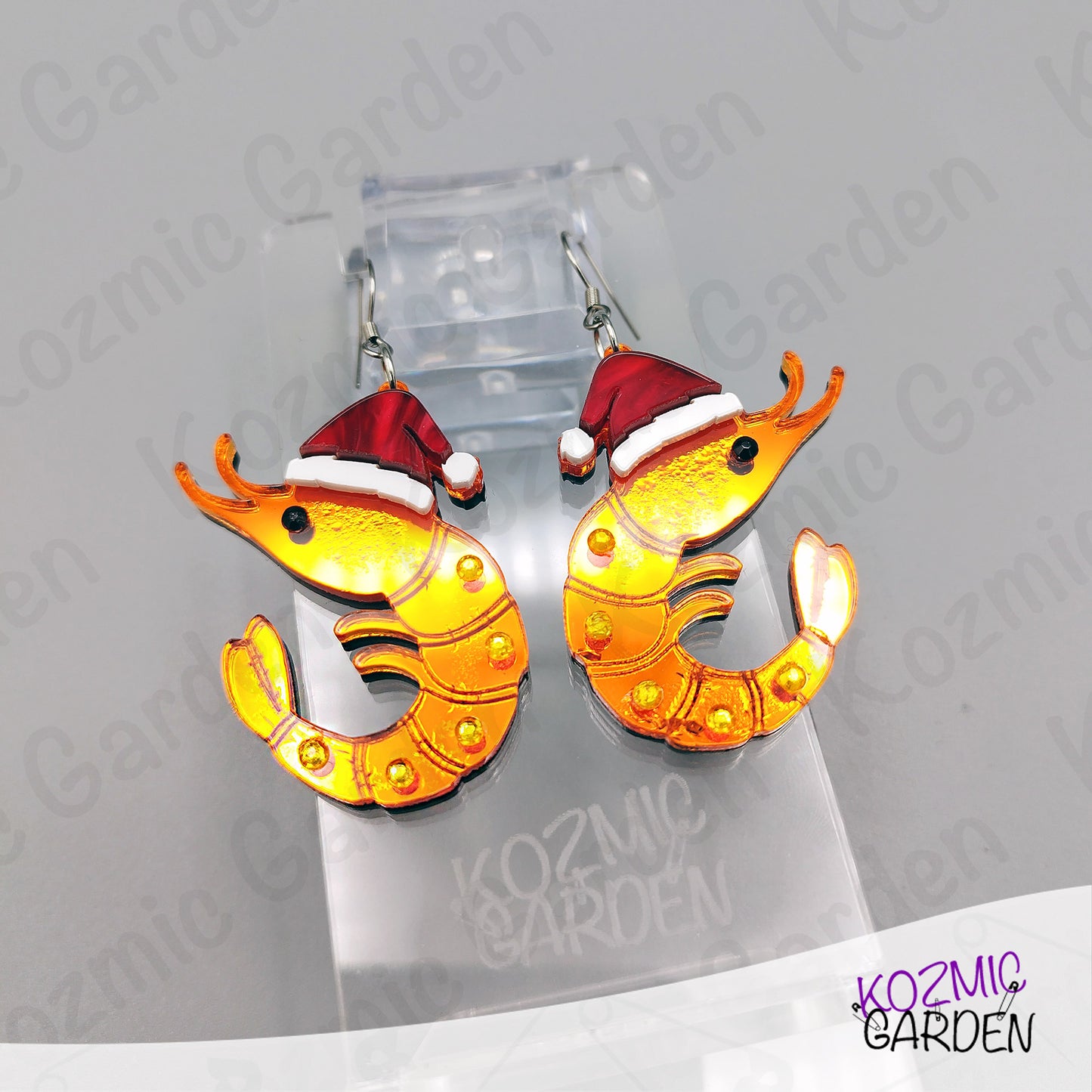 Shrimp-shaped earrings with Santa hats on a Kozmic Garden branded stand.