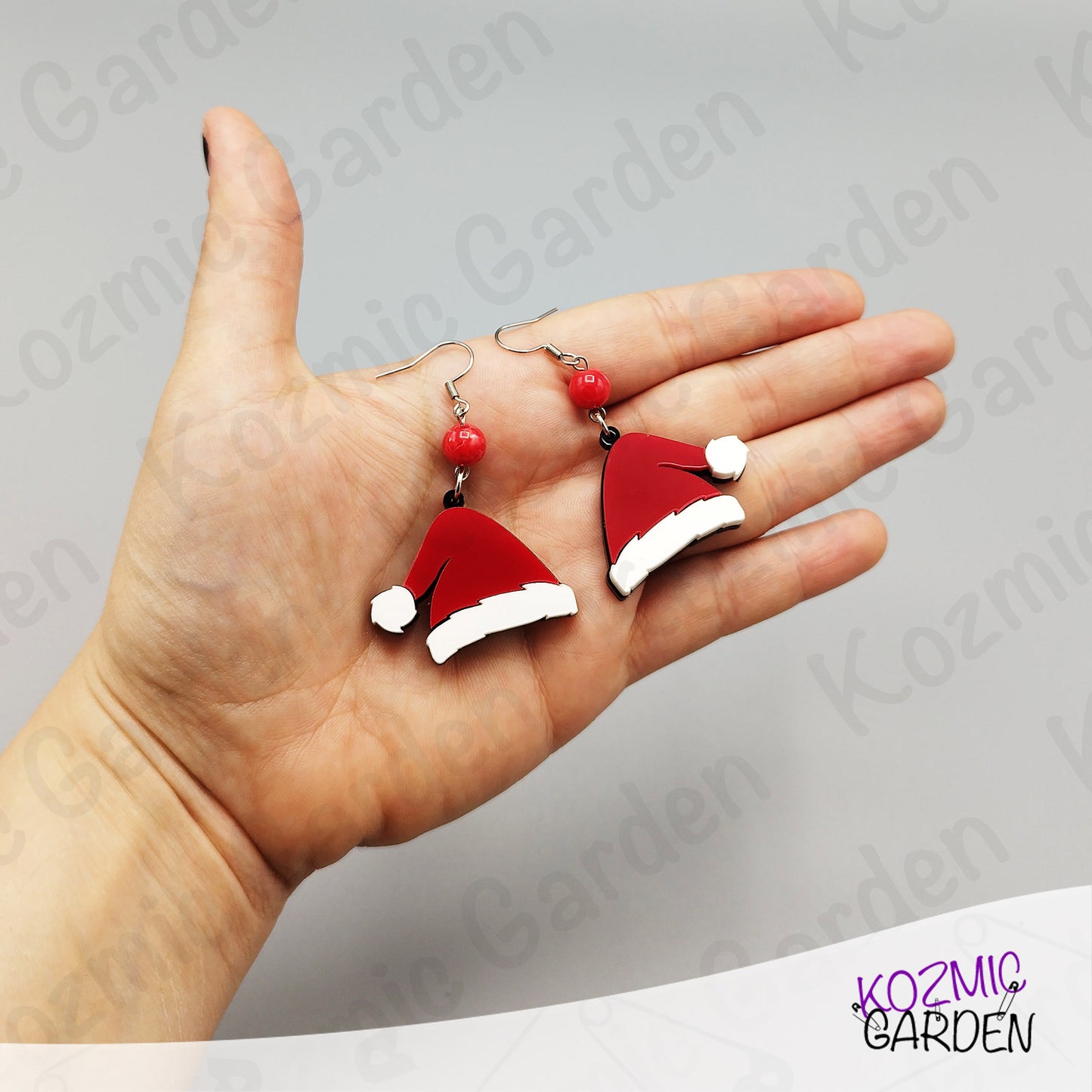 Hand holding Santa hat earrings with a gray background featuring 'Kozmic Garden' branding.